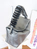 A dark gray pebbled leather handbag with a sculptural, twisted handle, displayed on a white stand against a white wall.