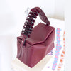 A structured burgundy leather handbag with a sculptural, layered handle, displayed on a white stand against a bright white background.