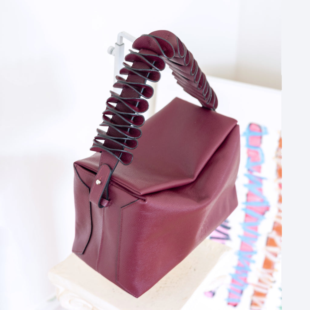 A structured burgundy leather handbag with a sculptural, layered handle, displayed on a white stand against a bright white background.