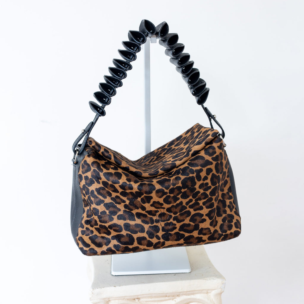 A leopard print shoulder bag with black accents and a coiled black handle, displayed on a white pedestal against a white background.