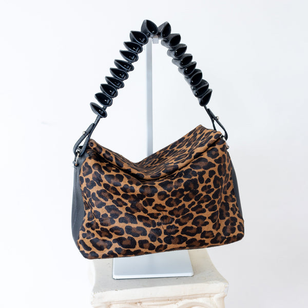 A leopard print shoulder bag with black accents and a coiled black handle, displayed on a white pedestal against a white background.