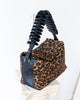A leopard print and black leather handbag with a sculptural spiral handle, displayed on a white pedestal against a white wall.