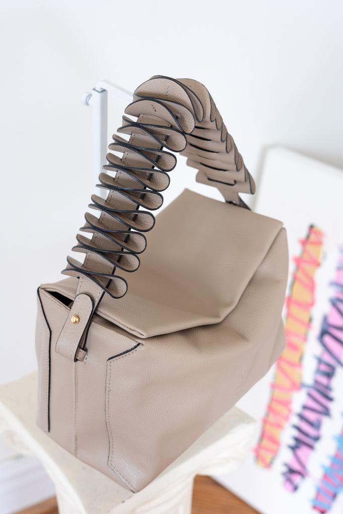 A structured taupe leather handbag with a sculptural, layered spiral handle, displayed on a white pedestal against a bright, minimalist background.