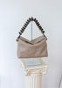 A taupe leather shoulder bag with a sculptural, coiled dark brown handle, displayed on a silver stand atop a white classical-style pedestal against a white wall.