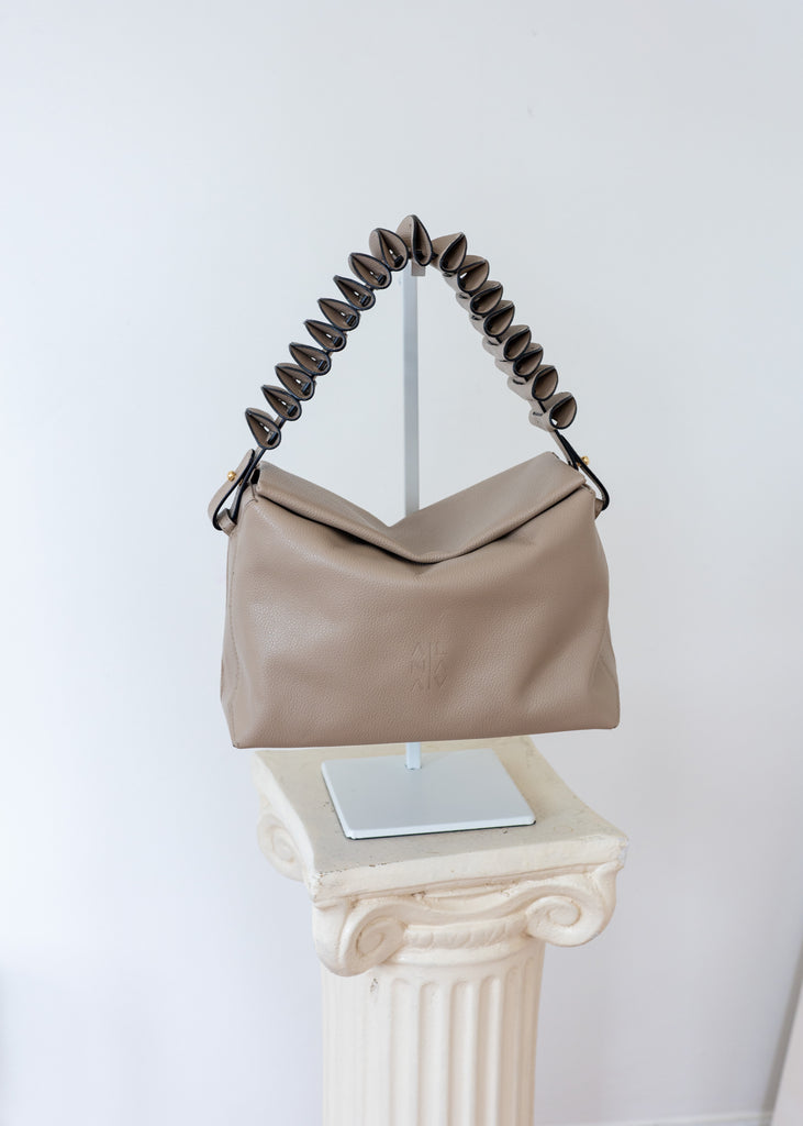 A taupe leather shoulder bag with a sculptural, coiled dark brown handle, displayed on a silver stand atop a white classical-style pedestal against a white wall.