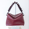 A burgundy pebbled leather shoulder bag with a sculptural, coiled dark red handle, displayed on a white stand over a stone pedestal against a white background.