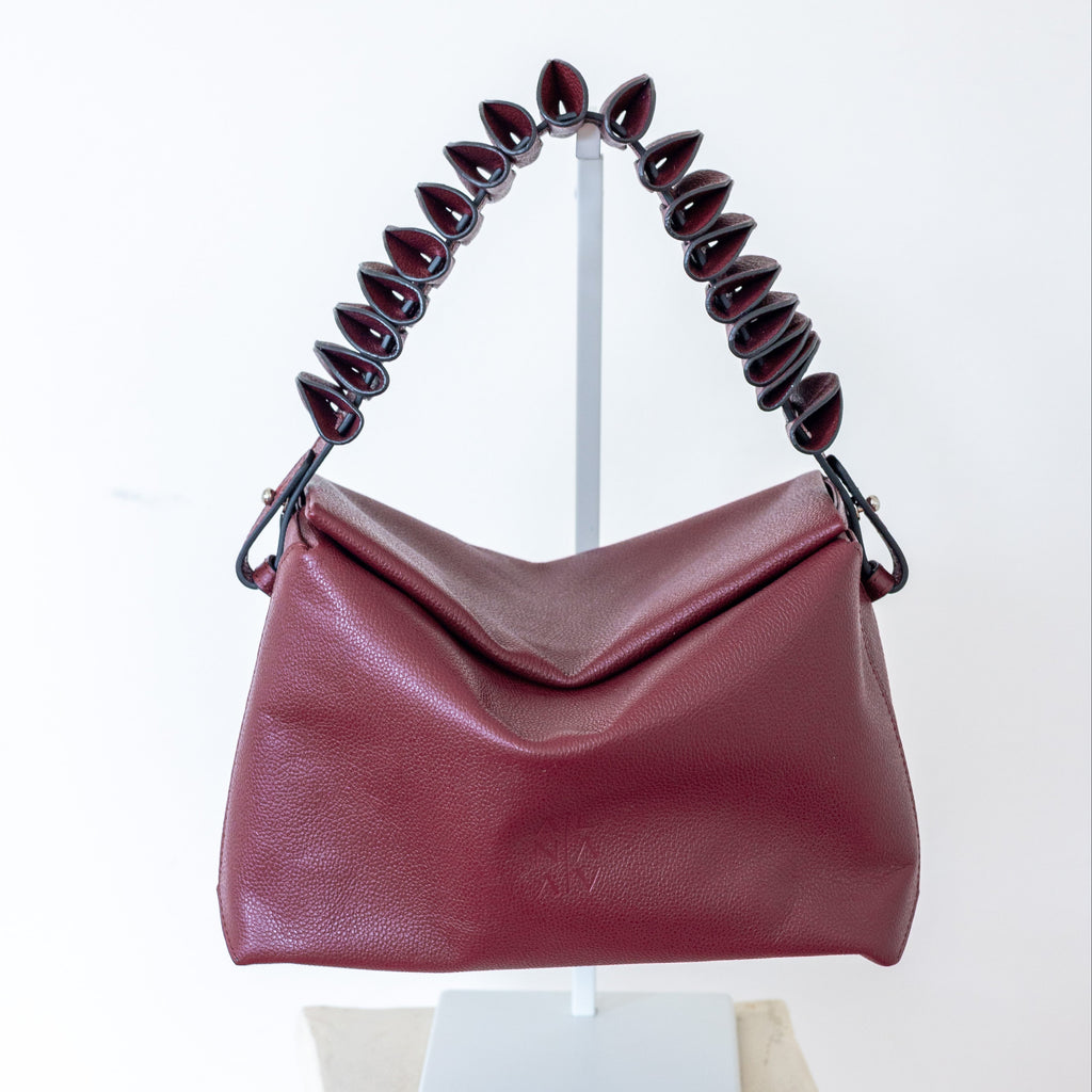 A burgundy pebbled leather shoulder bag with a sculptural, coiled dark red handle, displayed on a white stand over a stone pedestal against a white background.