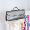 A metallic silver pebbled leather clutch handbag with a top handle, resting on a white decorative column against a light gray wall.