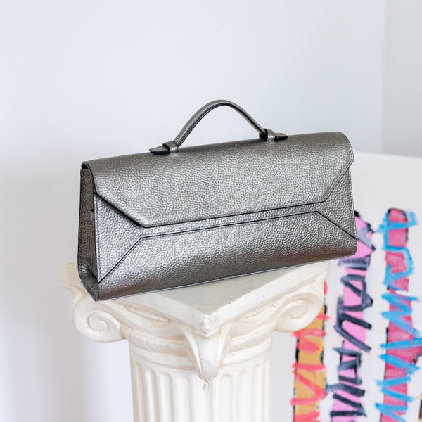 A metallic silver pebbled leather clutch handbag with a top handle, resting on a white decorative column against a light gray wall.