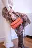 A person seated on a white column in an indoor room, wearing a cream long-sleeve top, a brown and beige patterned skirt, snakeskin knee-high boots, and holding a coral-orange envelope clutch.