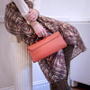 A person seated on a white column holds a structured coral-orange pebbled leather handbag with a top handle. The person wears a cream long-sleeve top, a brown and beige abstract print skirt, and snakeskin-patterned tights in an indoor setting.