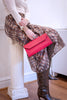 A person seated on a white column indoors, wearing a cream long-sleeve top, a brown and beige animal-print skirt, snakeskin knee-high boots, and holding a bright red envelope-style clutch.