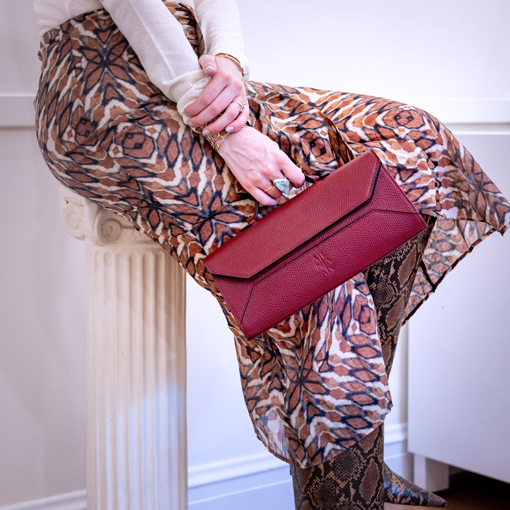 A person seated on a white column holding a structured burgundy leather clutch bag. The person wears a cream long-sleeve top, a brown and beige patterned skirt, and snakeskin boots in an indoor setting.