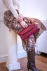A person seated on a white column wearing a cream long-sleeve top, a brown and beige floral print skirt, and snakeskin-print knee-high boots, holding a burgundy envelope-style clutch.