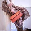 A person seated on a white column holds a structured orange envelope-style handbag. The person wears a brown and beige floral print skirt, a white long-sleeve top, a gold cuff bracelet, and snakeskin boots in an indoor setting.