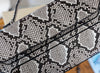 Close-up of a snakeskin-patterned handbag or clutch with black trim and a strap, shown against a softly blurred indoor background.