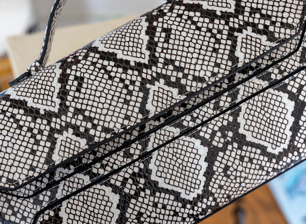 Close-up of a snakeskin-patterned handbag or clutch with black trim and a strap, shown against a softly blurred indoor background.