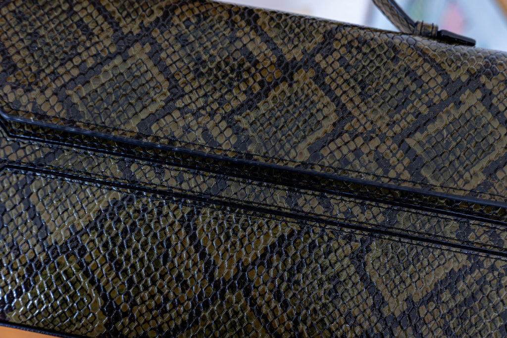 Close-up of a snakeskin-patterned handbag in olive green and black, showing the textured scale pattern and a black leather trim.