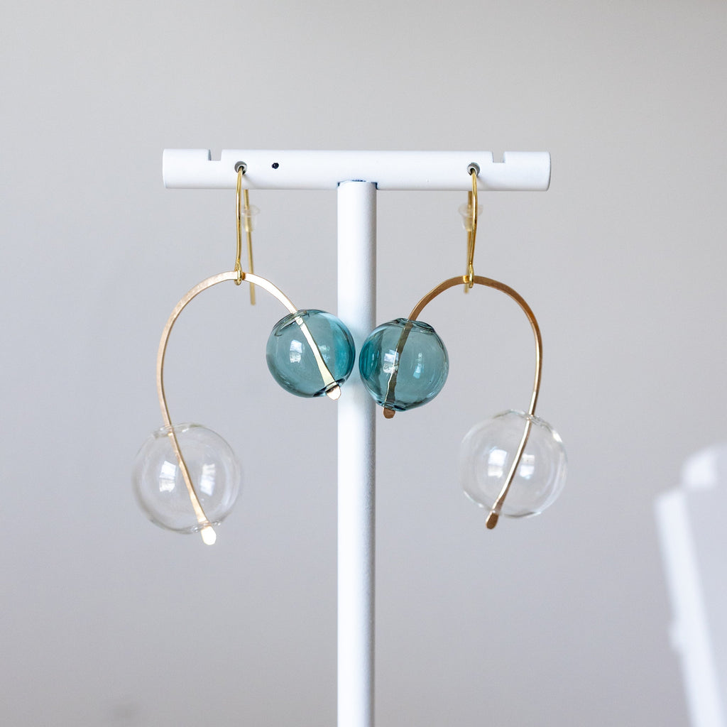 Earrings with gold loops and teal and clear glass beads on a white stand against a light gray background.