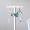 Earrings with gold loops and teal and clear glass beads on a white stand against a light gray background.