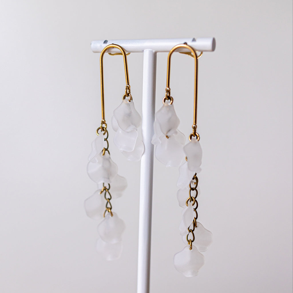 A pair of long dangle earrings with gold-toned metal hooks and translucent white flower-shaped beads, displayed on a white jewelry stand against a soft light gray background.