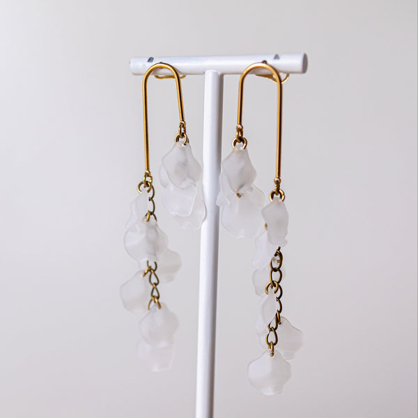 A pair of long dangle earrings with gold-toned metal hooks and translucent white flower-shaped beads, displayed on a white jewelry stand against a soft light gray background.