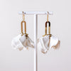 A pair of white sculptural dangle earrings with gold-toned metal connectors, hanging from a white display stand against a light neutral background.