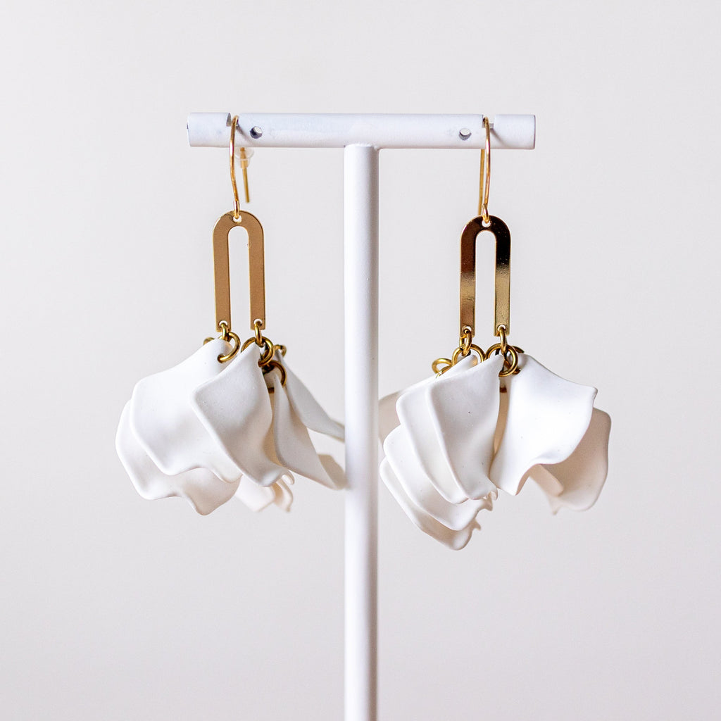 A pair of white sculptural dangle earrings with gold-toned metal connectors, hanging from a white display stand against a light neutral background.