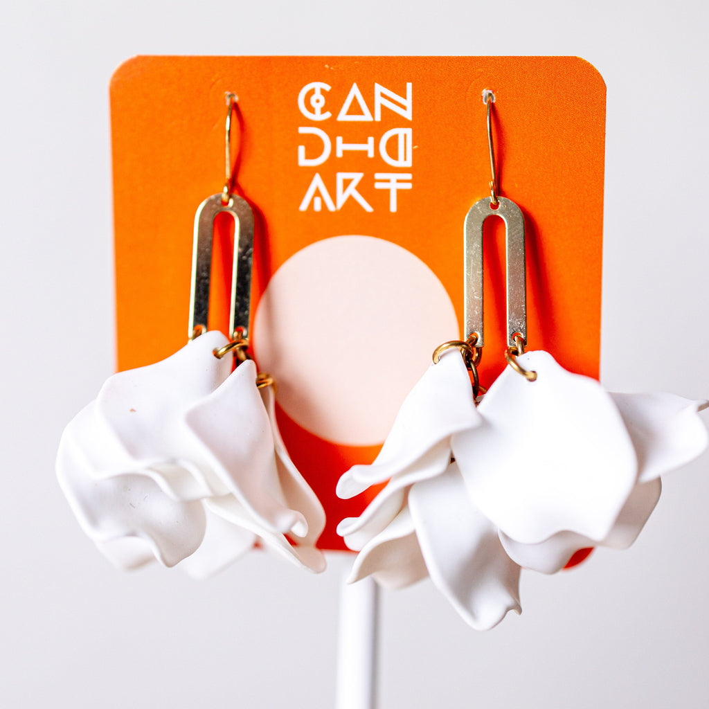 A pair of white sculptural flower dangle earrings with gold-toned metal hooks, displayed on an orange card that reads 'CANDID ART'.