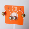 A pair of geometric hoop earrings with metallic gold and silver spheres, displayed on an orange card stand with the brand name 'CAN DO ART'.