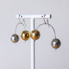 Three metallic ball earrings in silver and gold finishes hanging from a white display stand against a light gray background.