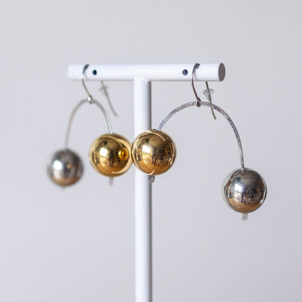 Three metallic ball earrings in silver and gold finishes hanging from a white display stand against a light gray background.