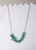 A gold-tone necklace with seven translucent light blue-green beads arranged in a V-shape, displayed against a white scalloped wooden background.
