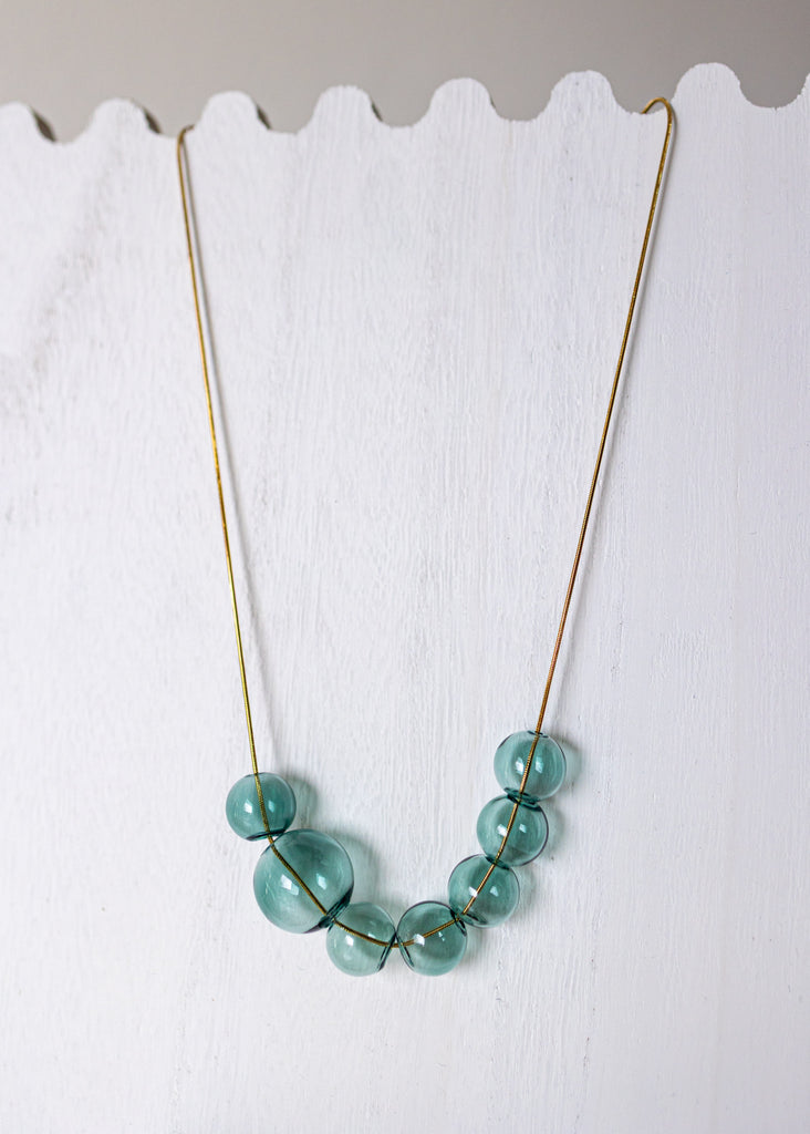 A gold-tone necklace with seven translucent light blue-green beads arranged in a V-shape, displayed against a white scalloped wooden background.