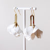 A pair of white sculptural dangle earrings with gold-toned hooks and rectangular metal connectors, displayed on a white stand against a soft light gray background.