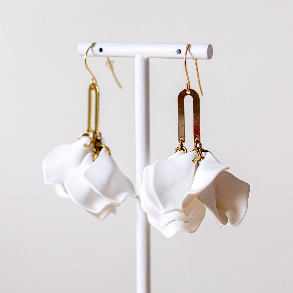 A pair of white sculptural dangle earrings with gold-toned hooks and rectangular metal connectors, displayed on a white stand against a soft light gray background.