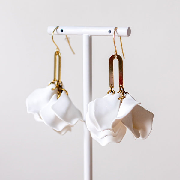 A pair of white sculptural dangle earrings with gold-toned hooks and rectangular metal connectors, displayed on a white stand against a soft light gray background.