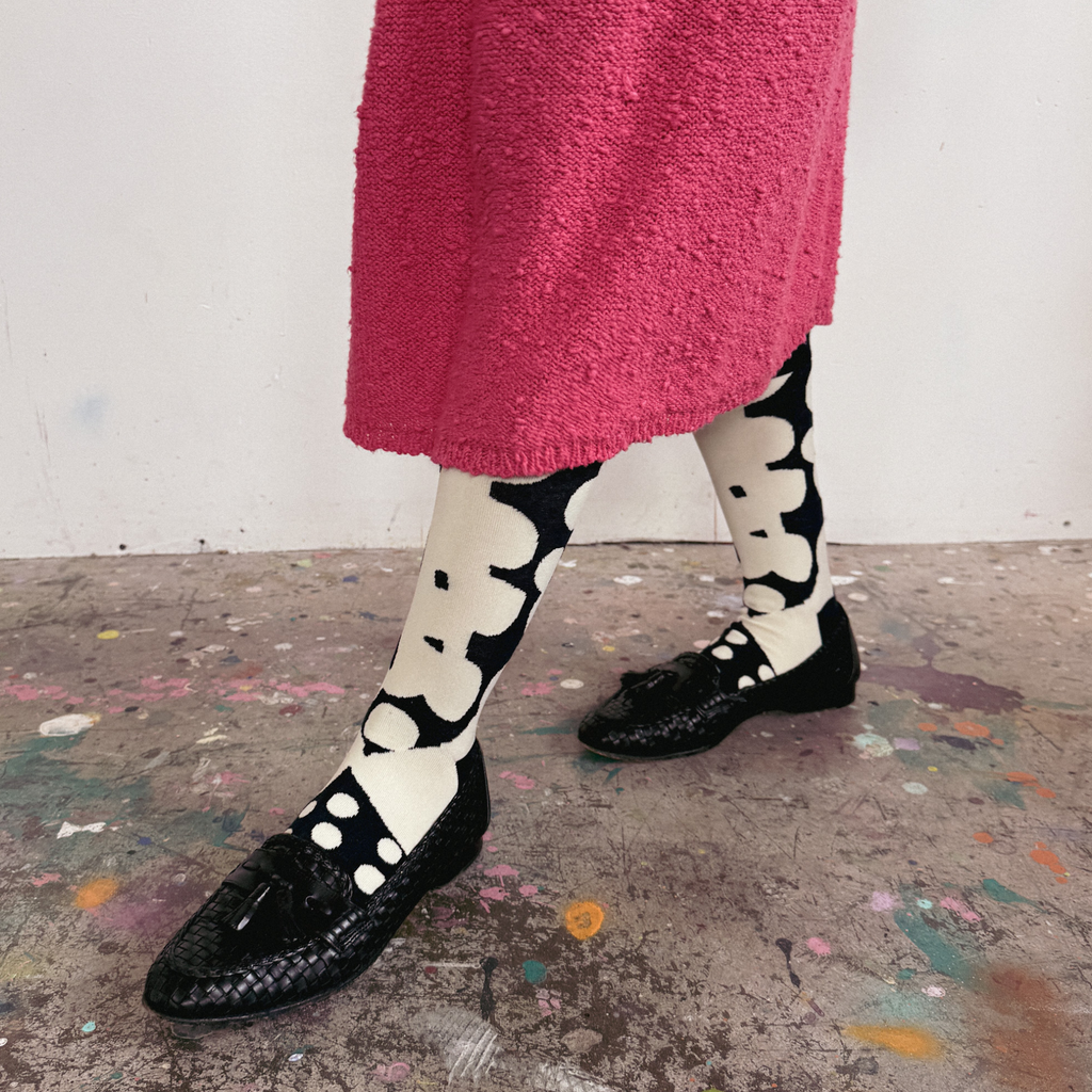 A person wearing black woven flat shoes with tassel details, black-and-white patterned socks, and a bright pink textured skirt, standing on a paint-splattered floor against a white wall.