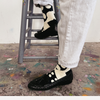 A person wearing black woven loafers and black-and-cream polka-dot socks, standing on a gray metal stool in a paint-splattered room.
