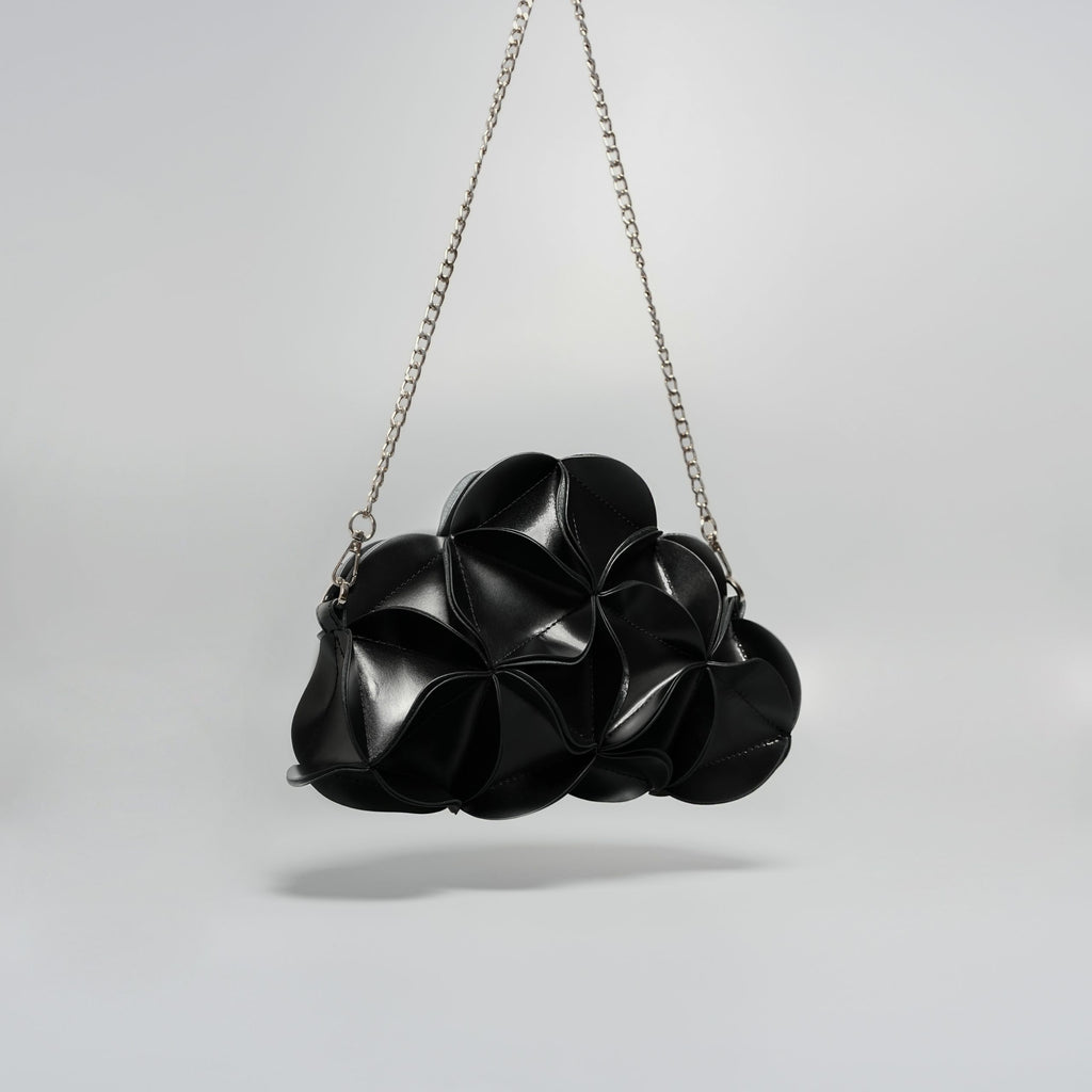 A black textured shoulder bag with a unique, organic, flower-like layered design and a silver chain strap, photographed against a neutral light gray background.