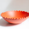 A bright coral-orange ceramic bowl with a scalloped rim, displayed on a white surface against a light gray background.