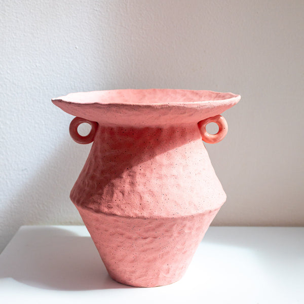 A textured terracotta pink ceramic vase with a wide, slightly flared rim and two small loop handles on the sides, displayed on a white surface against a neutral off-white wall.