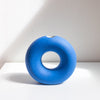 A bright blue donut-shaped ceramic vase with a small hole at the top, displayed on a white shelf against a textured off-white wall with soft shadows.