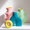 Four abstract ceramic vases in pink, teal, mint green, and yellow with speckled finishes, arranged on a white surface against a light beige wall.