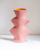 A sculptural, multi-tiered ceramic vase in a speckled dusty pink color, featuring a contrasting bright yellow-orange glazed interior, displayed on a white surface against a neutral light gray wall.