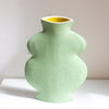 A light green speckled ceramic vase with a unique rounded, multi-bulb shape and a contrasting yellow interior, displayed on a white surface against a neutral beige wall.