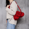 A person wearing a cream knit sweater and light blue jeans carries a bright red flower-shaped shoulder bag with a matching red strap against a gray concrete wall.