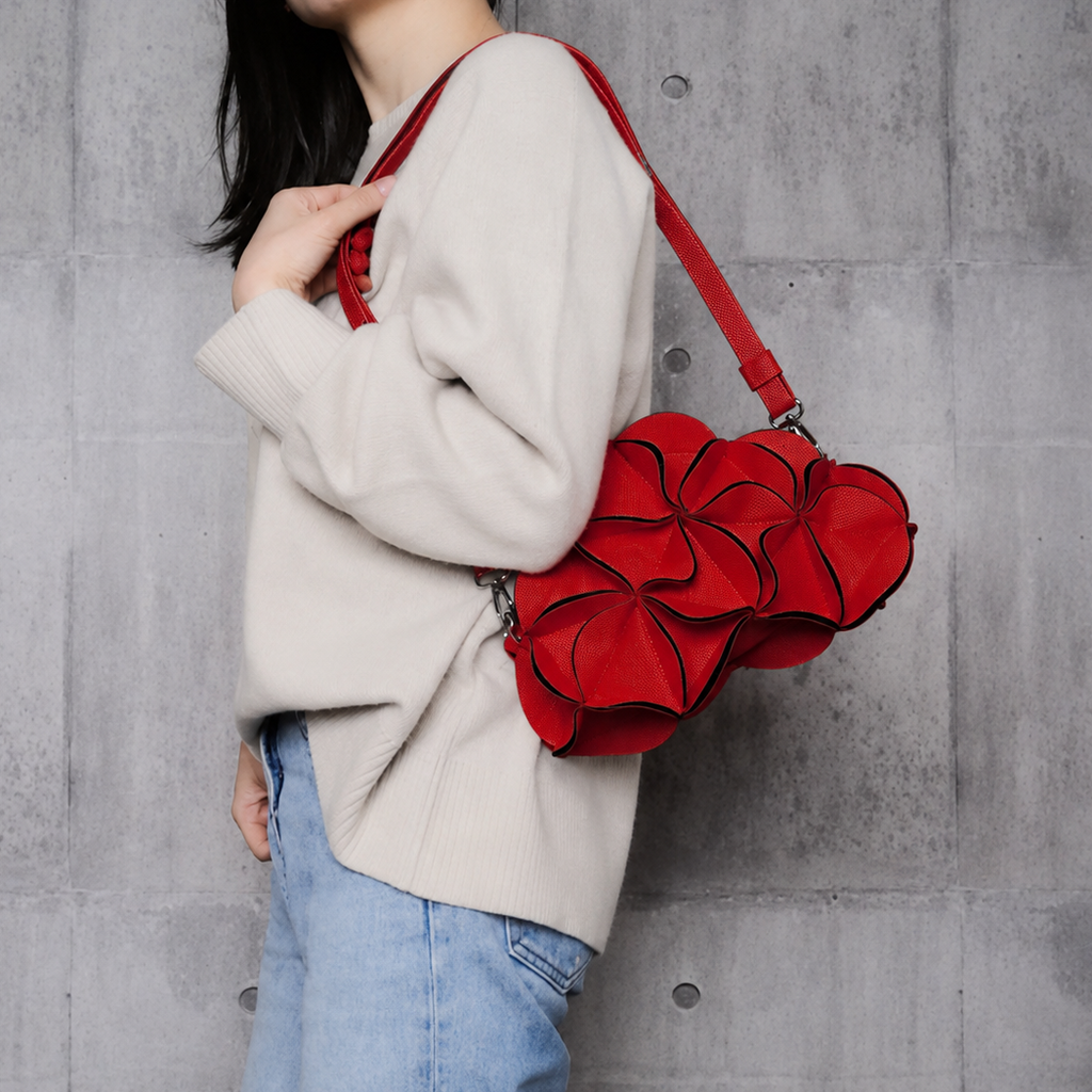 A person wearing a cream knit sweater and light blue jeans carries a bright red flower-shaped shoulder bag with a matching red strap against a gray concrete wall.