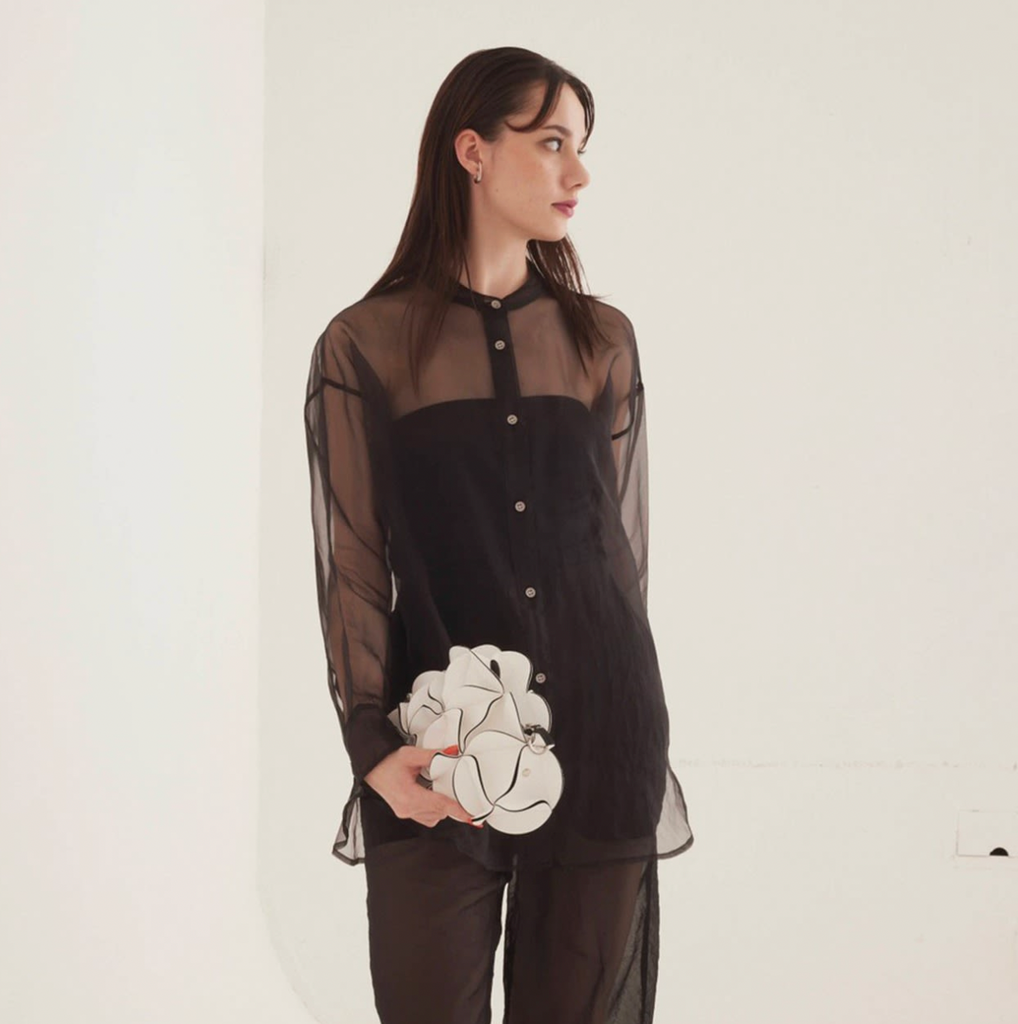 A woman stands in a minimalist white studio setting wearing a sheer black long-sleeve top with a structured black bodice and matching black trousers, holding a white sculptural clutch.