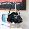 A black pebble-shaped shoulder bag with a sculptural, layered petal design, displayed on a silver stand in front of colorful abstract paintings.